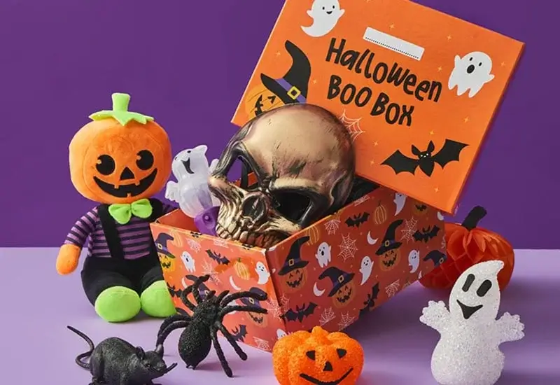 A Halloween-themed gift box labeled “Halloween Boo Box” filled with spooky items, including a metallic skull, small ghost figurines, and a black spider. Surrounding the box are festive decorations such as a plush pumpkin character, glittery ghosts, and mini pumpkins, all set against a purple background.